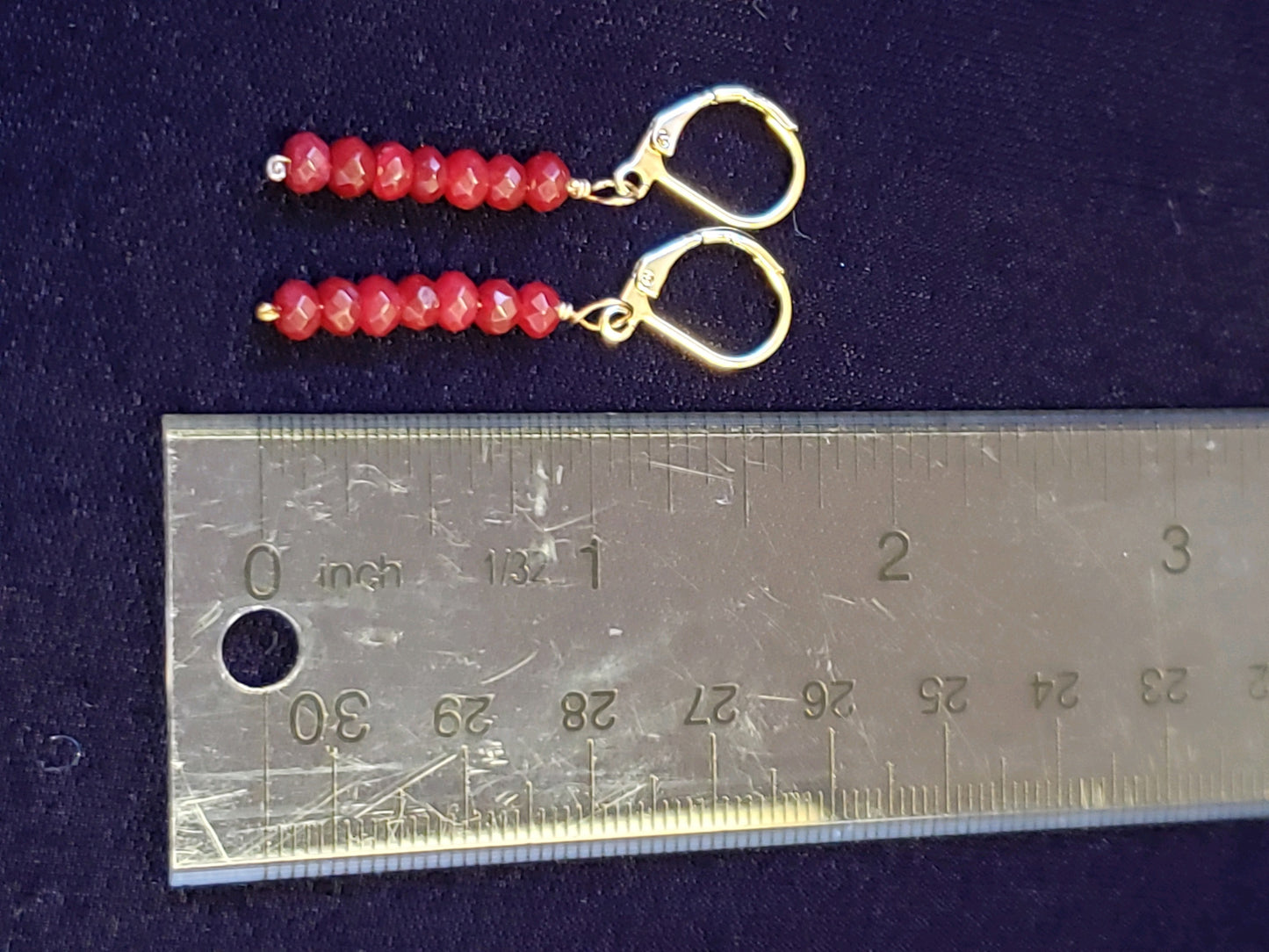 Pair of gold earrings with red beads next to a ruler on a dark background