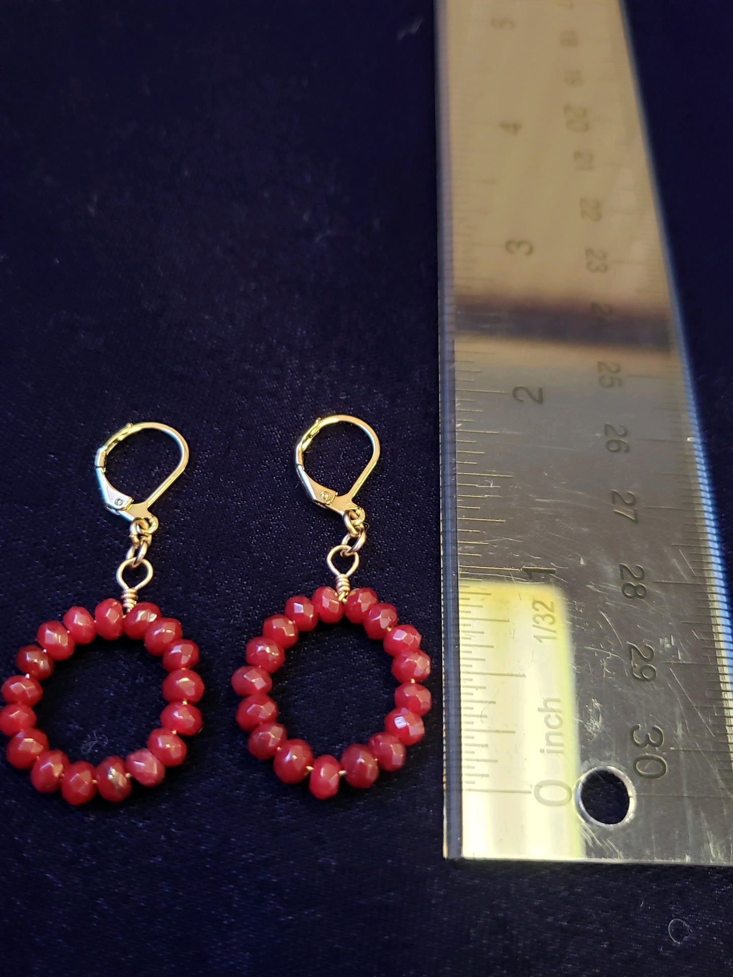 Pair of red beaded earrings with gold hooks on a dark background, next to a ruler for size reference.